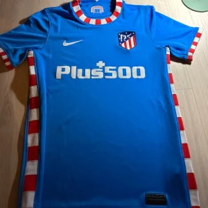 Atlético Madrid blå bortatröja Nike M - Säljer en blå Atlético Madrid bortatröja från Nike i Dri-FIT-material. Tröjan har korta ärmar och vita samt röda ränder på ärmslut, sidor och krage. Atlético-emblem på bröstet och Plus500-tryck framtill. Perfekt för dig som älskar fotboll och vill sticka ut.