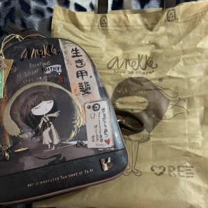 Anekke ryggsäck med konstnärligt motiv - Unik ryggsäck från Anekke med konstnärligt tryck och japanskinspirerade detaljer. Väskan är brun och svart med inslag av beige och guld, dekorerad med illustrationer, text och små berlocker. Tillverkad i syntetmaterial med flera fack och dragkedjor. Väskan är ny och äkta. 