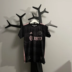 Inter Miami Messi matchtröja Adidas - Säljer en svart Inter Miami fotbollströja från Adidas med rosa detaljer och Messi 10 på ryggen. Tröjan har korta ärmar, klubbmärke på bröstet och sponsortryck framtill. Tillverkad i lätt och ventilerande polyester, perfekt för fotbollsplanen.