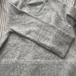 Grå stickad v-ringad tröja - Mysig grå stickad tröja med v-ringning och långa ärmar. Tröjan har breda ribbade muddar vid ärmslut och nederkant, vilket ger en snygg och stilren look. Perfekt för lager-på-lager under kyliga dagar. köpte för 279kr