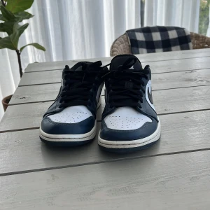 Nike Air Jordan 1 Low marinblå/vit - Säljer ett par Nike Air Jordan 1 Low i marinblått och vitt med svarta detaljer. Skorna har klassisk låg siluett, svart swoosh och snörning. Ovandelen är i skinn och sulan är vit med marinblå yttersula. Perfekta för dig som gillar streetwear och sneakers med ikonisk design.