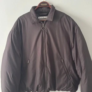 Svart pufferjacka från Acne Studios - Säljer en svart pufferjacka från Acne Studios med oversized passform. Jackan har en hög krage, två snedställda fickor med dragkedja och en stor rund dragkedjepull. Materialet är syntet och jackan är helfärgad utan mönster.