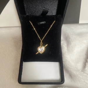 Guldigt halsband med diamant hjärthänge - Superfint halsband i guldton med ett gnistrande hjärthänge och pil-detalj. Hänget har en klar sten i mitten som ger extra bling. Perfekt accessoar för dig som gillar söta och romantiska smycken med lite edge. Rostfri 