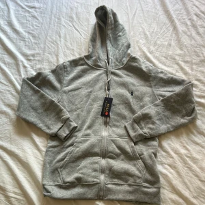 Grå hoodie från Polo Ralph Lauren - Säljer en grå hoodie från Polo Ralph Lauren med dragkedja och huva. Tröjan har snörning i huvan, två fickor fram och den klassiska broderade loggan på bröstet. Perfekt för chill dagar och enkel att matcha med jeans eller joggers.