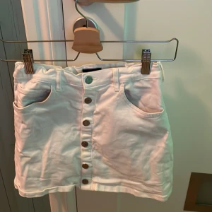 Vit jeanskjol från Gant med knappar - Snygg vit jeanskjol från Gant med knappar hela vägen framtill. Kjolen har klassiska fickor både fram och bak samt bälteshällor. Perfekt för dig som gillar en enkel men stilren look.