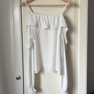 Vit volangblus från Gina Tricot - Supersöt vit blus från Gina Tricot med tunna axelband och bred volang längs urringningen. Långa, luftiga ärmar med resår och volangdetalj vid handlederna. Perfekt för en fräsch och romantisk look.