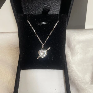 Halsband med hjärthänge i silver - Snyggt halsband i silverfärg med ett hjärtformat hänge som har en glittrande sten i mitten och en pil genom hjärtat. Kedjan är tunn och stilren, perfekt för att ge din outfit lite extra kärleksvibe. Passar dig som gillar romantiska detaljer och bling. Rostfri kjedjan är 40cm lång 💘💘