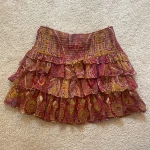 Färgglad volangkjol med paisleymönster - Supersöt kort volangkjol med paisleyinspirerat mönster i rött, gult och rosa. Kjolen har flera lager volanger och bred resår i midjan för en bekväm passform. Perfekt för dig som gillar boho-vibbar och färg! Materialet känns lätt och luftigt.