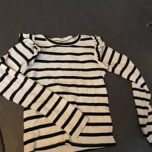Randig långärmad tröja med volang - Supersöt långärmad tröja med svarta och vita ränder och volangdetaljer på axlarna. Perfekt för dig som gillar en klassisk men ändå lekfull stil. Mjuk och skön, passar till jeans eller kjol för en chill look.
