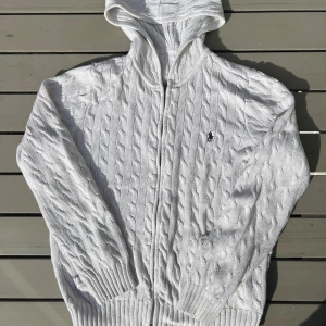 Vit kabelstickad hoodie från Ralph Lauren - Vit kabelstickad hoodie från Ralph Lauren i 100% bomull. Tröjan har dragkedja framtill, huva och ribbade muddar. Klassisk broderad logga på bröstet. Perfekt för dig som gillar en clean och stilren look.