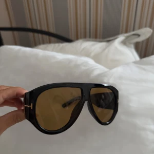 Svarta Tom Ford pilotbågar solglasögon - Snygga svarta solglasögon från Tom Ford med pilotbågar och bruna tonade glas. Ramen är i blank plast och har guldiga T-detaljer på sidorna. Perfekt accessoar för en cool look och passar till många outfits.
