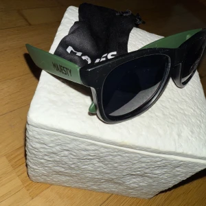 Svarta och gröna solglasögon Majesty - Snygga solglasögon från Majesty med svart front och gröna skalmar. Modellen har en klassisk wayfarer-form och mörka linser som ger cool vibe. Perfekt accessoar för att lyfta din stil och skydda ögonen mot solen.