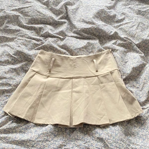 Beige plisserad kortkjol - Superfin beige kortkjol med plisserad design och bred linning. Kjolen har hällor i midjan och är sydd i ett mjukt material som känns bekvämt. Perfekt för dig som gillar en stilren och enkel look.