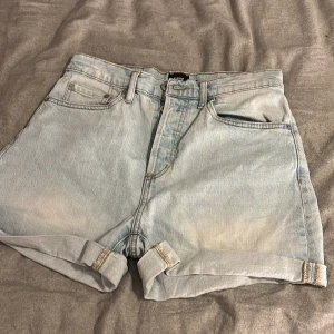 Ljusblå jeansshorts från 157 - Snygga ljusblå jeansshorts från 157 med klassisk femficksdesign och uppvikta benslut. Modellen är högmidjad och har slitna detaljer för en chill vibe. Perfekta för varma dagar och enkel att matcha med din favorit-tshirt eller hoodie.
