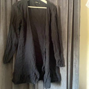 Svart lång klänning från BikBok XS - Svart lång klänning från BikBok i storlek XS. Klänningen har långa ärmar, volangdetalj nedtill och är öppen framtill utan knäppning. Tillverkad i en skön blandning av linne och viskos, perfekt för lager-på-lager-stil.