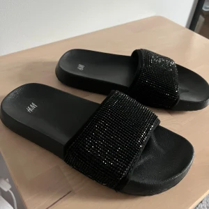 Svarta glittriga sandaler från H&M - Svarta sandaler från H&M med bred rem täckt av små svarta glittrande pärlor. Platt sula med räfflad undersida för extra grepp. Perfekt om man ska utomlands eller som innetofflor. Väldigt bekväma och aldrig använda, endast testade.