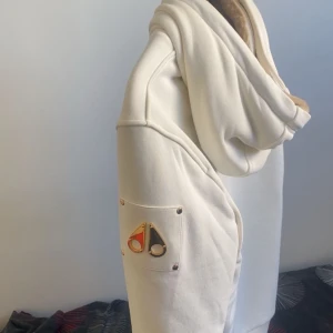 Vit hoodie från Moose Knuckles - Säljer en vit hoodie från Moose Knuckles med guldfärgade detaljer på ärmen och svart dragkedja. Insidan har quiltad svart foder och beige fuskpäls längs huvan. Tröjan har snörning vid huvan och två fickor framtill. Perfekt för kalla dagar. Har haft den på mig ba 2 gånger