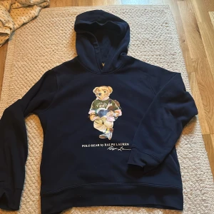Mörkblå Polo Bear hoodie från Ralph Lauren - Säljer en mörkblå hoodie från Ralph Lauren med ikoniska Polo Bear-trycket på bröstet. Tröjan har huva och långa ärmar, tillverkad i mjuk bomullsmix. Perfekt för dig som vill ha en klassisk och snygg streetstyle-look.