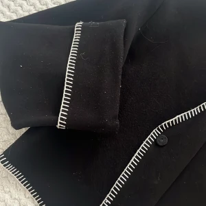 Svart kappa från Gina Tricot med kontrastsöm - Svart kappa från Gina Tricot med snygga vita kontrastsömmar längs kanterna. Jackan har en klassisk krage, knäppning framtill och långa ärmar. Perfekt för dig som gillar stilrena plagg med en twist.