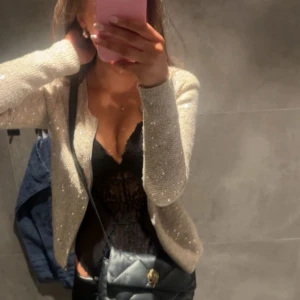 Beige glittrig kofta från Flash Woman City - Snygg beige kofta med glittriga paljetter från Flash Woman City. Koftan har lång ärm, rund hals och dragkedja framtill. Materialet är stickat och ger en mysig känsla, perfekt för att lyfta din outfit med lite extra bling.