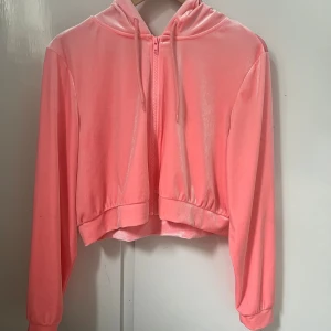 Rosa croppad hoodie från Monki - Säljer en supermjuk och glansig rosa hoodie från Monki i croppad modell. Tröjan har dragkedja framtill, huva med snören och långa ärmar. Perfekt för dig som gillar en chill och trendig stil.