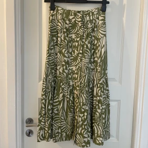 Suncoo Grönmönstrad långkjol  - Superfin långkjol från Suncoo Paris med grönt och vitt tropiskt mönster. Kjolen har knappar framtill och är sydd i ett luftigt linnetyg, perfekt för varma dagar. Den har en lätt volang nedtill som ger extra flow och en snygg siluett.