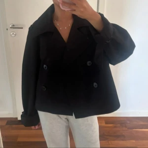 Svart trenchcoat med knappar - Kort svart trenchcoat med dubbelknäppning och bred krage. Jackan har långa ärmar med uppvikta muddar och en loose passform som ger en chill och trendig look. Perfekt för dig som gillar klassisk stil med modern twist.