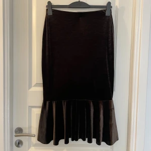 Svart sammetskjol med volang från Paulmark - Supersnygg svart sammetskjol från Paulmark i storlek 38. Kjolen är lång och har en volang nertill som ger en cool och trendig look. Perfekt för dig som gillar stilrena plagg med en twist. Materialet är mjukt och glansigt, vilket gör kjolen extra lyxig.