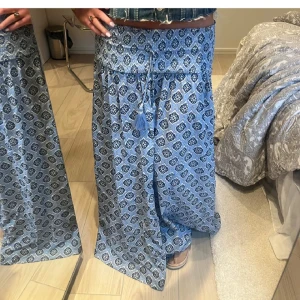 Blå långkjol med mönster och snörning - Säljer en blå långkjol med snyggt grafiskt mönster och justerbar snörning i midjan med tofsar. Kjolen har en avslappnad passform och är perfekt för dig som gillar boho-stil. Lätt och luftig, passar grymt till sneakers eller sandaler. I stlrk 158 164