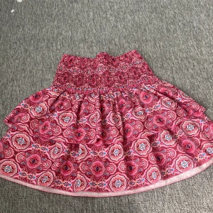 Rosa mönstrad volangkjol - Supersöt kort kjol med bred smock i midjan och volanger. Kjolen har ett färgstarkt mönster i rosa, rött, blått och vitt. Använd en gång