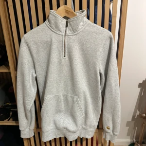 Carhartt ljusgrå sweatshirt, storlek XS  - Ljusgrå sweatshirt med hög krage och dragkedja. Sweatshirten har en ficka på magen och en Carhartt-logga på ärmen.