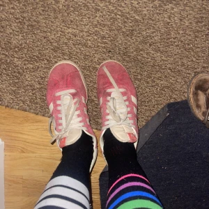 Adidas Gazelle röda sneakers mocka - Säljer ett par klassiska Adidas Gazelle sneakers i röd mocka med vita detaljer och snörning. Skorna har de ikoniska tre ränderna på sidan och texten 'GAZELLE' i guld. Platt sula och rund tå, perfekt för en sportig och retro look. Byter man snöret och gnuggar lite med vatten ser dem som nya ut! Köpt för 1300 PRIS KAN DISKUTERAS