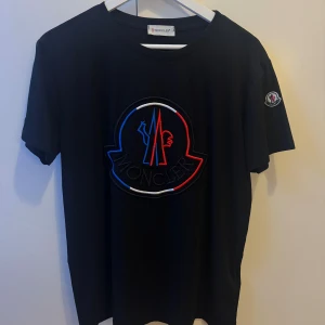 Svart Moncler t-shirt med stor logga - Svart t-shirt från Moncler i bomull med stor broderad logga i blått, rött och vitt på bröstet. Klassisk passform med korta ärmar och rund hals. Moncler-märke även på ena ärmen. Perfekt för dig som gillar streetwear och exklusiva märken. Den är i väldigt bra skick då jag använt den bara 1 gång. Det är en 1️⃣av 1️⃣, hög kvalitet 