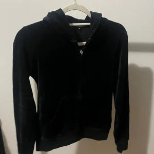 Y2K Juicy Couture svart velour hoodie - Y2K kofta Juicy Couture hoodie i svart velour med dragkedja framtill och huva. Baksidan har en stor broderad krona i guld och blått samt texten 'Viva La Glam'. Perfekt statement-plagg för dig som gillar glam och streetstyle.