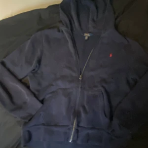 Marinblå hoodie från Polo Ralph Lauren - Säljer en marinblå hoodie från Polo Ralph Lauren med röd broderad logga på bröstet. Tröjan har dragkedja, huva och känguruficka. Perfekt för dig som gillar klassisk och stilren hoodies
