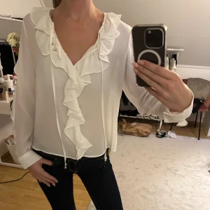 Vit blus med volangdetaljer - Supersöt vit blus med volang längs v-ringningen och ärmarna. Lätt och luftig känsla, perfekt för dig som gillar romantisk stil. Blusen har långa ärmar och knytband framtill. Passar lika bra till jeans som till kjol. Nästan aldrig använd💕