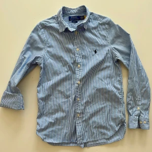 Randig skjorta från Polo Ralph Lauren - Snygg blå- och vitrandig skjorta från Polo Ralph Lauren med klassisk krage och knappar framtill. Skjortan har lång ärm och ikonisk broderad logga på bröstet. Perfekt för en stilren look.
