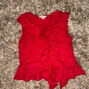 Röd volangblus från Gina Tricot - Supersöt röd blus från Gina Tricot i storlek 146/152. Blusen är ärmlös med volangdetaljer längs halsringningen och framtill. Materialet är lätt skrynklat och består av bomull, polyester och elastan för en bekväm känsla.