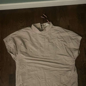 Beige topp med hög krage Holly & Whyte - Säljer en beige topp från Holly & Whyte i storlek XS. Toppen har hög krage med snygg öppning och knäppning bak samt en dekorativ randig rosett. Modellen är ärmlös och har en rak, loose passform. Perfekt för dig som gillar stilrena och enkla plagg.