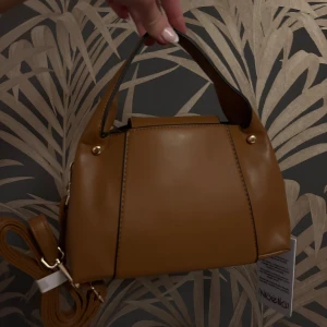 Brun handväska från Noella - Snygg brun handväska från Noella i veganskt skinn. Väskan har ett stilrent och modernt utseende med guldfärgade detaljer och både handtag och avtagbar axelrem. Perfekt storlek för det viktigaste och passar till många outfits. I nytt skick med prislapp kvar! 