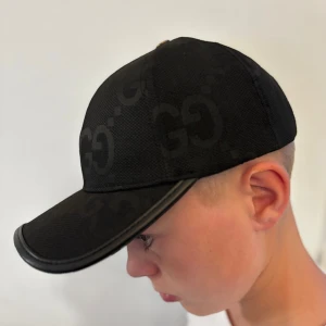 Svart keps med GG-mönster - Snygg svart keps  med diskret GG-mönster över hela kepsen. Böjd skärm med svart kant och justerbar rem baktill i svart med gulddetalj. Perfekt för dig som gillar exklusiva accessoarer med stilren design.