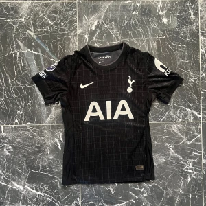 Tottenham svart matchtröja Bergvall 15 - Snygg svart Tottenham Hotspur fotbollströja från Nike med Bergvall och nummer 15 på ryggen. Tröjan har korta ärmar, diskret rutmönster och vita tryck med AIA-logga på bröstet samt Premier League-märke på ärmen. Materialet är lätt och andas, perfekt för match eller träning.