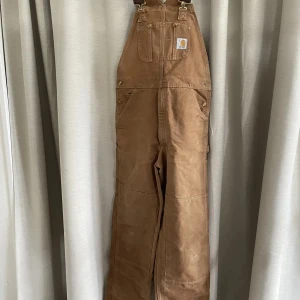 Bruna Carhartt hängselbyxor - Säljer ett par klassiska bruna hängselbyxor från Carhartt med flera praktiska fickor och robusta detaljer. Byxorna har breda axelband, förstärkta knän och är tillverkade i slitstark bomullscanvas.  