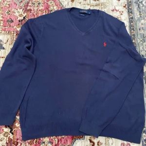 Marinblå v-ringad tröja Polo Ralph Lauren - Snygg marinblå v-ringad tröja från Polo Ralph Lauren i mjuk pima bomull. Klassisk design med röd broderad logga på bröstet och ribbade muddar vid ärmslut och nederkant. 