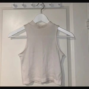 Beige ribbad ärmlös polotopp - Säljer en beige, croppad polotopp i ribbad bomull. Toppen är ärmlös med hög hals och har en clean, minimalistisk look som passar perfekt till jeans eller kjol. Enkel att styla och superbekväm för varmare dagar.