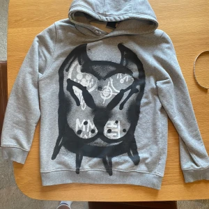 Grå Givenchy hoodie med svart print - Säljer en grå hoodie från Givenchy med ett stort svart och vitt grafiskt print framtill. Tröjan har huva, långa ärmar och en relaxed passform. Givenchy-logga på ryggen. Perfekt för dig som gillar streetwear och vill sticka ut. I tröjan står det 12+ men är endast lite liten för mig som är 178 cm. Skriv och fråga om du funderar över något, t.ex. passformen!