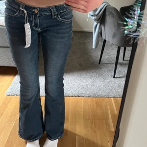 Blå bootcut jeans med broderade fickor - Snygga mörkblå bootcut jeans och roderade bakfickor i beige och guld. Jeansen har låg midja och klassisk femficksdesign. Perfekta för dig som gillar western-inspirerad stil.