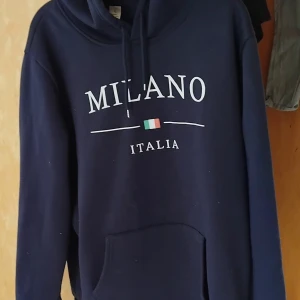 Mörkblå Milano hoodie med huva - Snygg mörkblå hoodie med trycket 'MILANO ITALIA' och en liten italiensk flagga på bröstet. Tröjan har huva med snörning och en stor magficka. Perfekt för chill dagar och streetstyle. Passar dig som gillar enkel och stilren design.