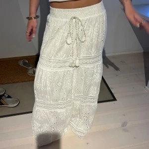 Vit broderad långkjol med snörning - Superfin vit långkjol med broderade blommor och snygga spetsdetaljer. Kjolen har resår i midjan och en dekorativ snörning med tofsar framtill. Perfekt för sommarens alla tillfällen och ger en riktigt fräsch och bohemisk vibe.