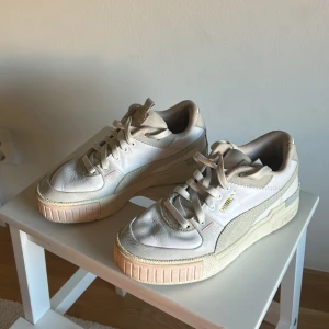 Beige och vita sneakers från Puma - Snygga sneakers från Puma i vitt och beige med detaljer i mocka och skinn. Skorna har klassisk snörning, rund tå och platt sula. Guldiga Puma-loggor på sidan och hälen ger en extra touch. Perfekta för dig som gillar stilrena och neutrala färger.